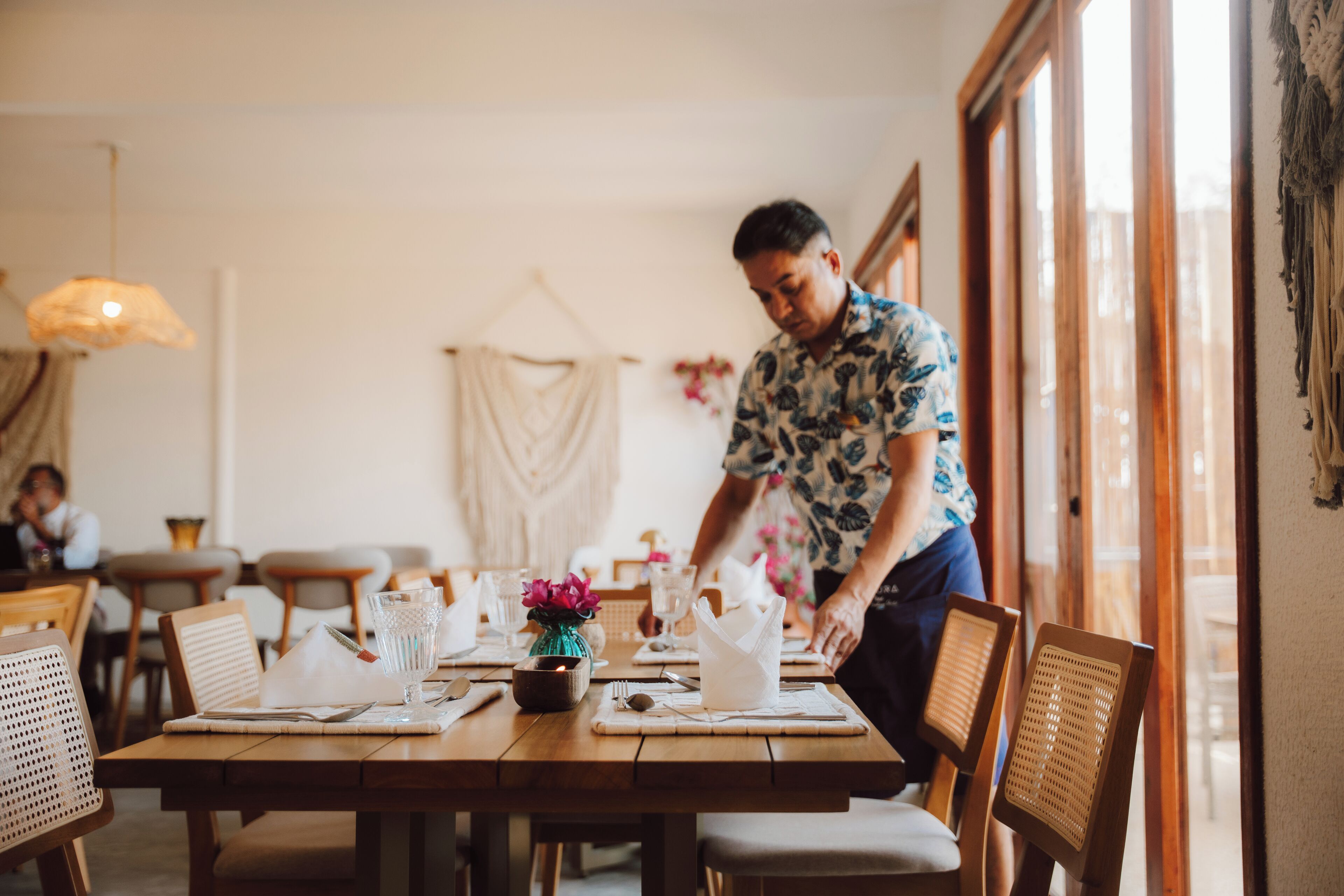 Un comedor con mesas y sillas de madera, un jarrón con flores, y una persona preparando la mesa.