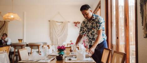 Un comedor con mesas y sillas de madera, un jarrón con flores, y una persona preparando la mesa.