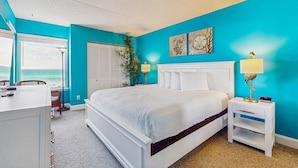 A bedroom with a large bed, white nightstands, a desk, and a view of the ocean. - Comfortable beachfront condo with beach setup - quick drive to dining! (Fort Walton Beach)