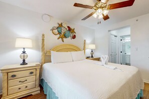 A neatly made bed with white bedding, a decorative wall art piece, a ceiling fan, and a nightstand with a lamp. - Cozy oceanfront condo with ocean view, pool access, washer&dryer, and central AC (Fort Walton Beach)