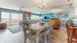 A spacious living area with a wooden dining table, chairs, and a sofa. There is a balcony with a view, a kitchen area, and a dining bar. - Second floor oceanfront condo with ocean view, balcony, central AC, & pool access (Fort Walton Beach)
