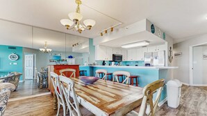 A spacious living area with a wooden dining table, white chairs, and a kitchen with white cabinets and a blue accent wall. - Second floor oceanfront condo with ocean view, balcony, central AC, & pool access (Fort Walton Beach)