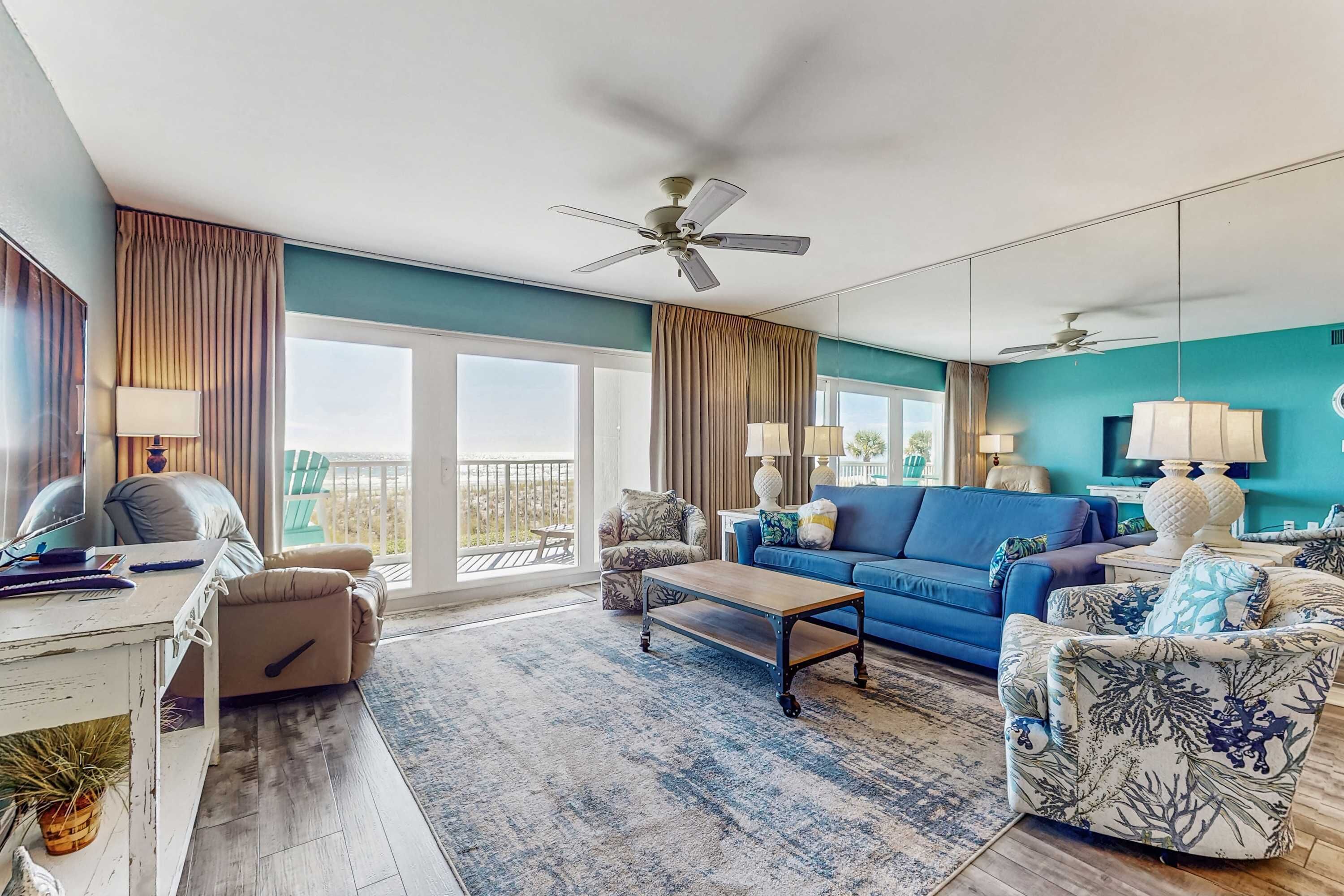A spacious living room with a blue sofa, a coffee table, and a patterned armchair. There is a large window with curtains, a ceiling fan, and a balcony visible through the window.