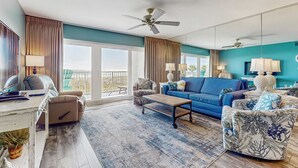 A spacious living room with a blue sofa, a coffee table, and a patterned armchair. There is a large window with curtains, a ceiling fan, and a balcony visible through the window. - Second floor oceanfront condo with ocean view, balcony, central AC, & pool access (Fort Walton Beach)