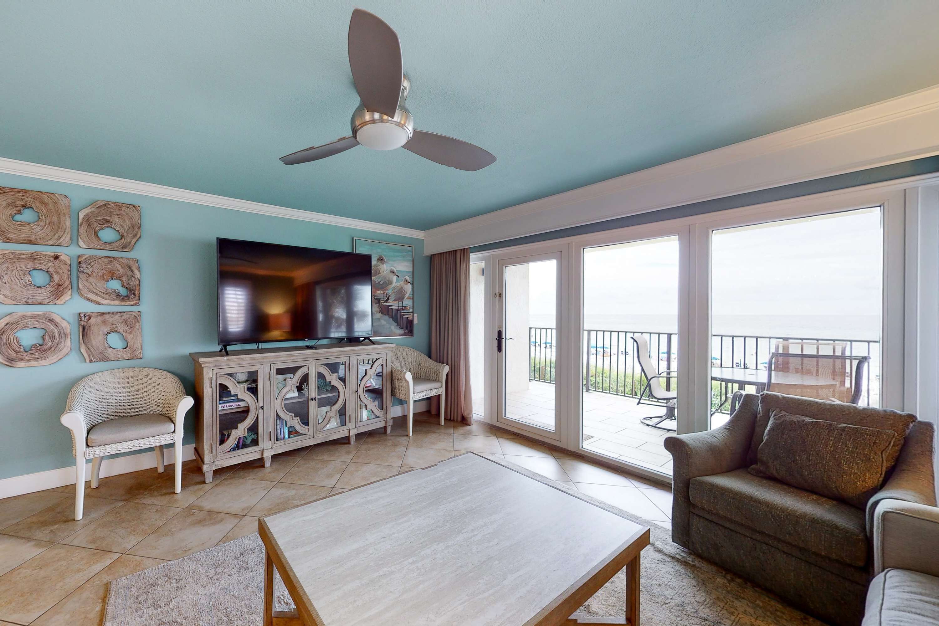 Una sala de estar con ventilador de techo, televisor, sofá, mesa de centro y balcón con mobiliario exterior.