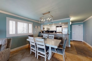 A dining area with a wooden table and chairs, a chandelier, and a kitchen in the background. - Gorgeous beachfront condo with a pool & private beach access (Miramar Beach)
