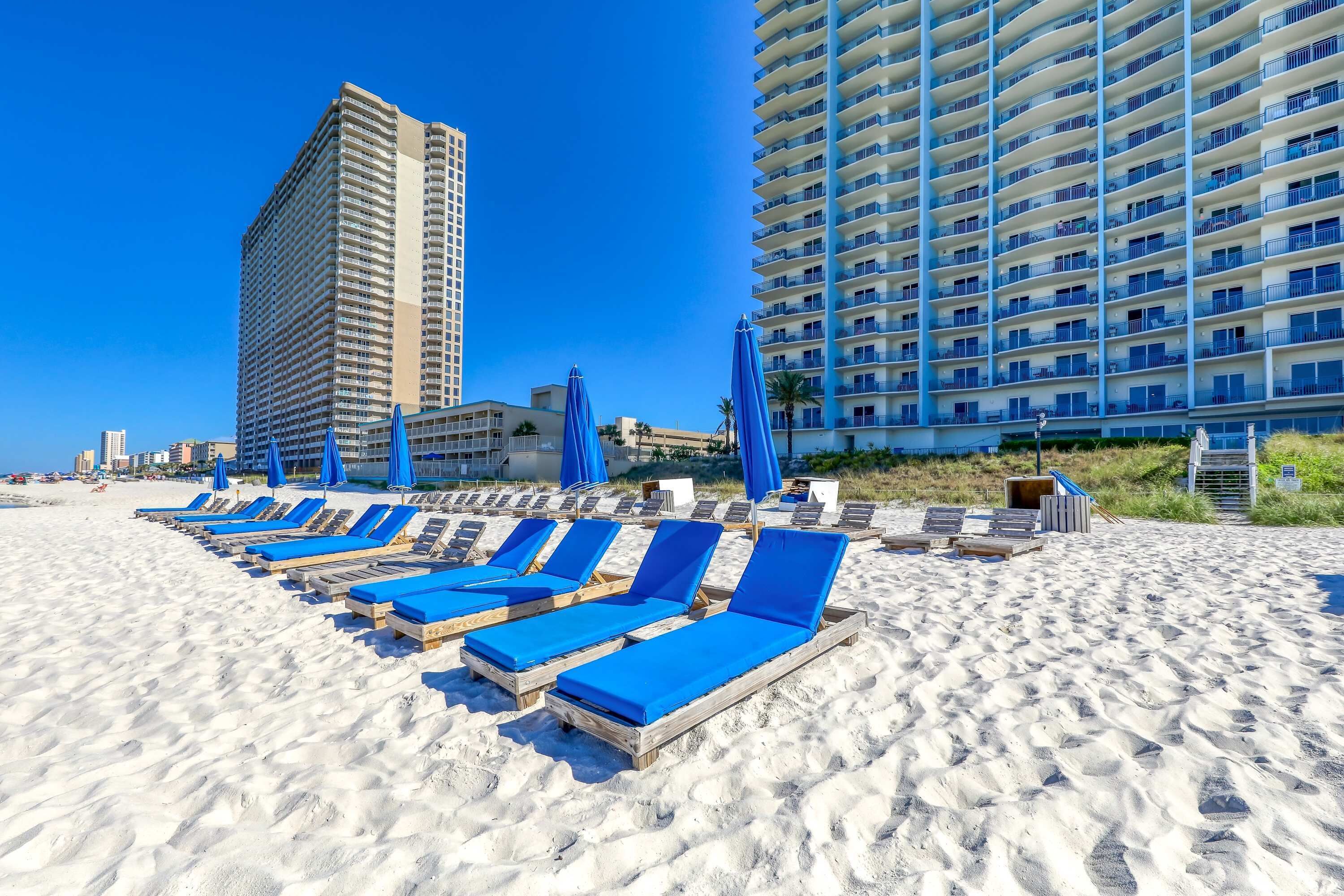 A beach with sun loungers and umbrellas, a tall building, and a hotel with balconies.
