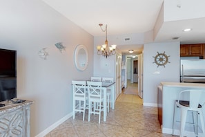 A modern kitchen with white cabinetry, a stainless steel refrigerator, and a dining area with white chairs and a table. - Flat with stunning view, pool, & beach access (Panama City Beach)