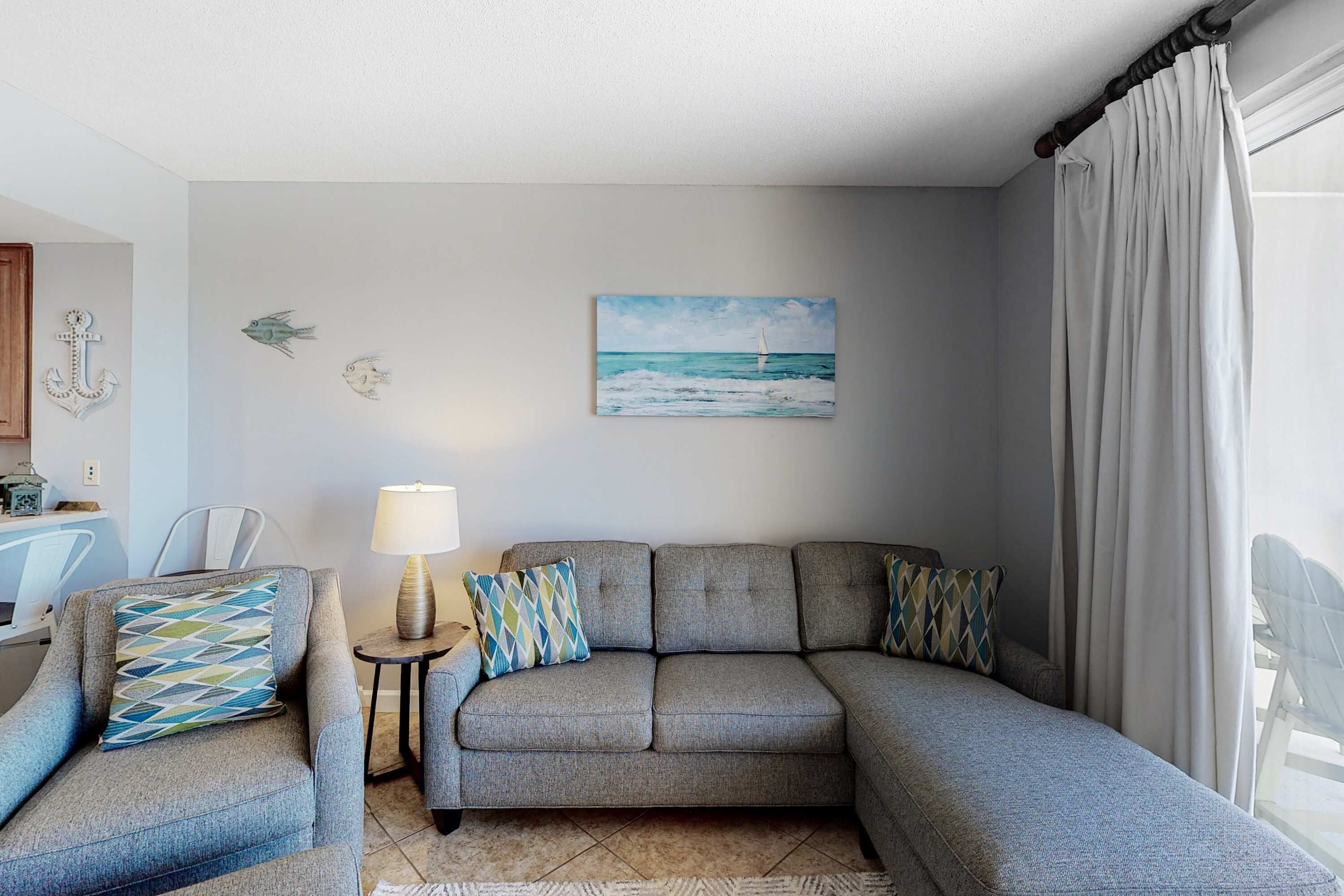 Un salon avec un canapé gris, des coussins à motifs, une petite table avec une lampe et un tableau représentant un voilier accroché au mur.