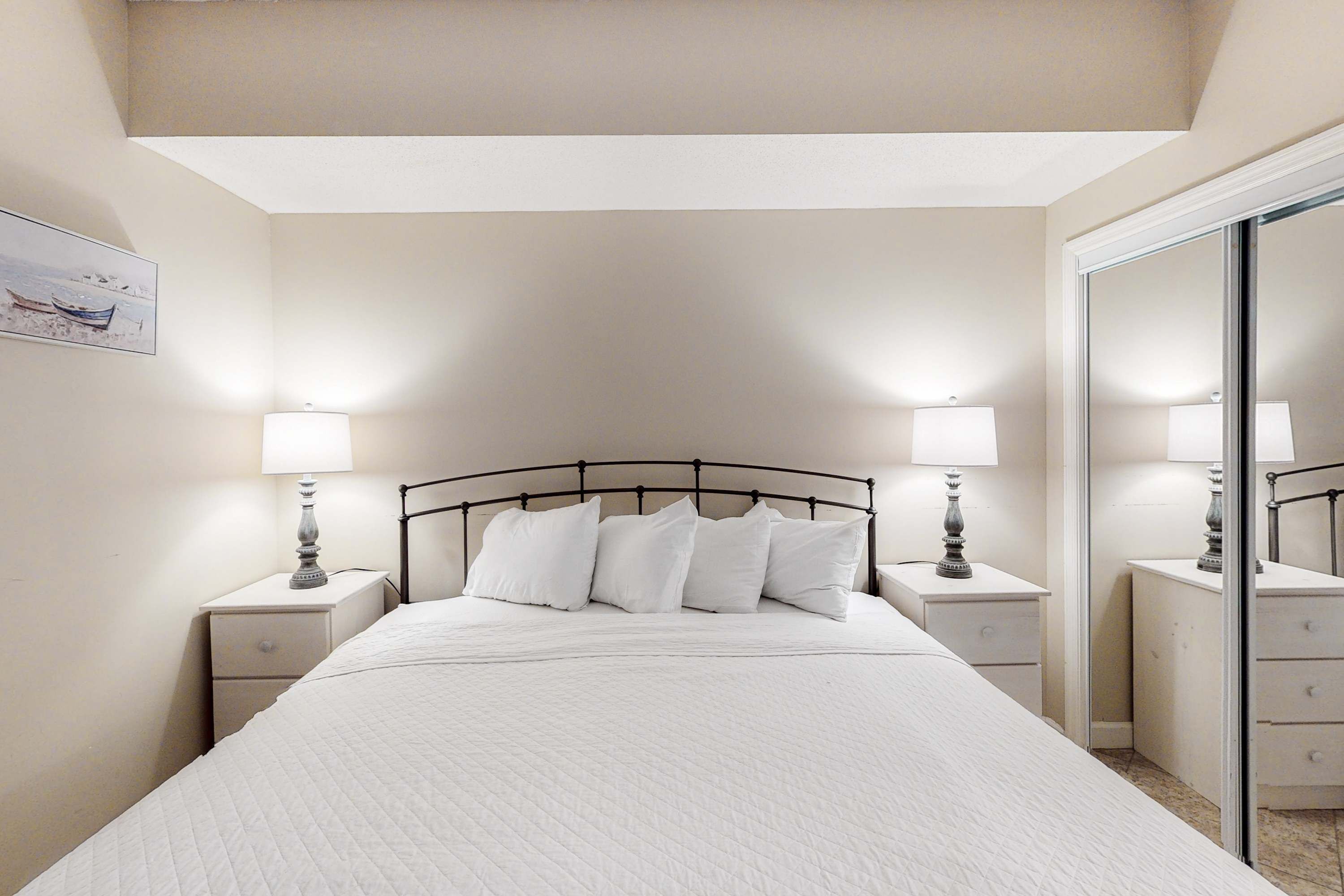 A neatly arranged bedroom with a large bed, two bedside tables with lamps, a mirror, and a painting on the wall.