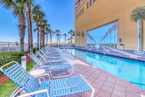 A poolside area with lounge chairs and a swimming pool, surrounded by palm trees and a large building with a distinctive archway. - Gulf-front condo with free beach service, balcony & pool (Panama City Beach)
