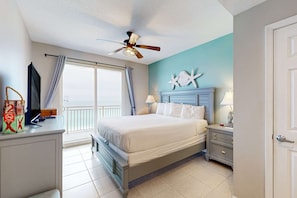 A bedroom with a bed, a nightstand, a dresser, a ceiling fan, a window with curtains, and a wall decoration. - Gulf-front condo with free beach service, balcony & pool (Panama City Beach)