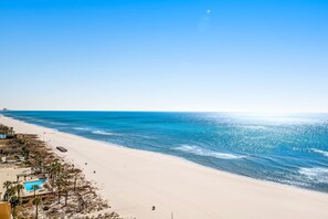 A beach with a clear sky, a sandy shore, and a small pool area with umbrellas. - Gulf-front condo with free beach service, balcony & pool (Panama City Beach)