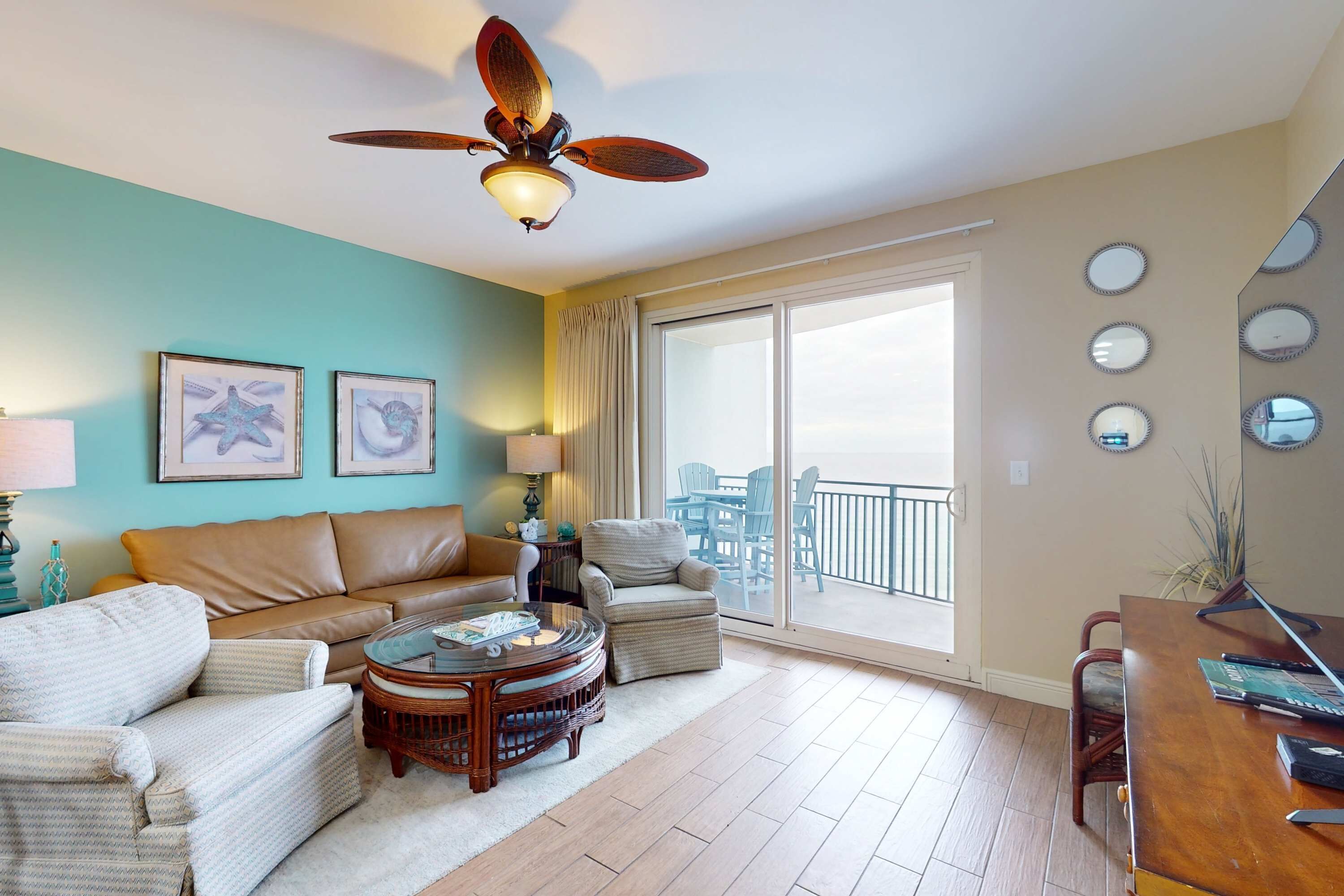 Un salon avec un ventilateur de plafond, un canapé, une table basse et une table à manger. Les murs sont ornés de tableaux encadrés et il y a une grande fenêtre offrant une belle vue.