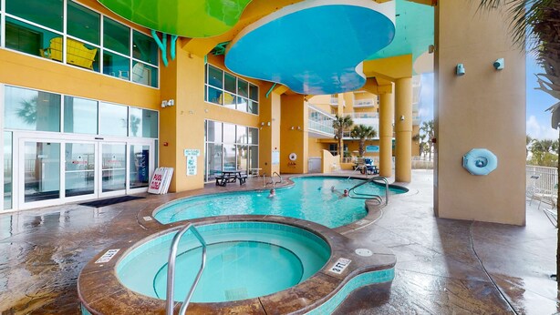 Uma área de piscina com várias piscinas, um espaço coberto com assentos e um edifício com grandes janelas e fachada amarela.