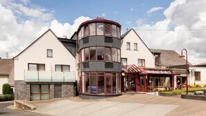 A modern building with a curved glass tower, a covered outdoor seating area, and a paved entrance. - Landhotel Wolf-Mertes (Sessenbach)
