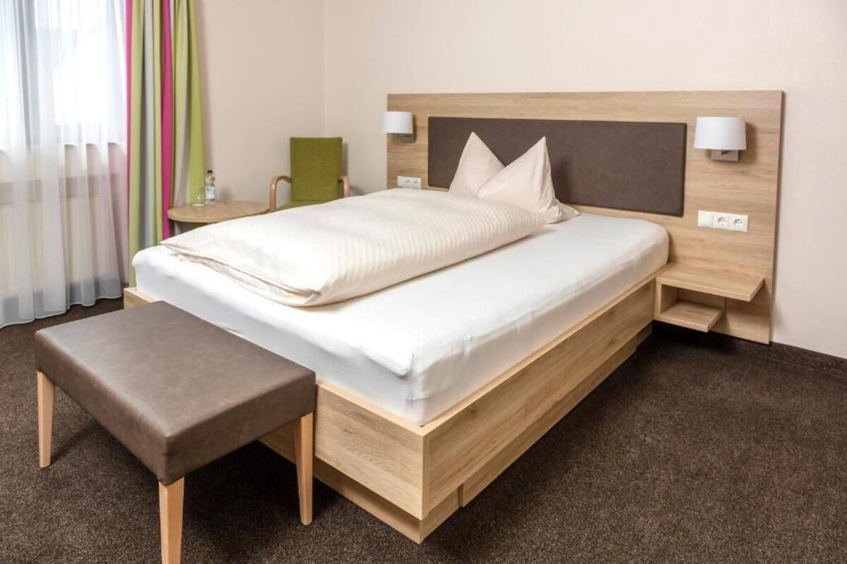 A hotel room with a wooden bed, a bench, two bedside tables, and a window with sheer curtains.