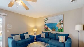Un salon avec un canapé bleu, une table basse en verre, un tableau représentant une scène de plage et un ventilateur de plafond.
