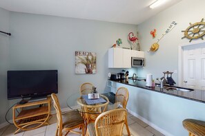 Ein kompakter Wohnbereich mit Essbereich, ausgestattet mit einem Glastisch und Korbstühlen, einer Küchenzeile inklusive Mikrowelle und Toaster sowie einem Fernseher auf einem Schrank.