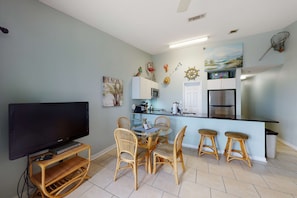 Een compacte keuken met een eethoek, met een flatscreen-tv, rieten stoelen en een klein keukeneiland met barstoelen.