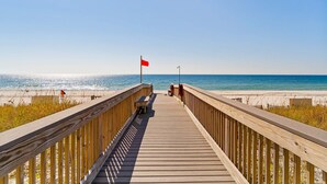 A wooden boardwalk leading to a beach with a red flag, a bench, and a lamp post. - Gorgeous Oceanfront condo with washer/dryer, Gulf-front balcony & pool/hot tub (Perdido Key)