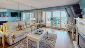 A modern living room with a beige sofa, a flat-screen TV, and a balcony with outdoor furniture. - Gorgeous Oceanfront condo with washer/dryer, Gulf-front balcony & pool/hot tub (Perdido Key)