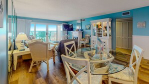 A room with a glass dining table surrounded by white chairs, a sofa, and a TV. There are windows with curtains, a cabinet with various items, and a door with a sign. - Gorgeous Oceanfront condo with washer/dryer, Gulf-front balcony & pool/hot tub (Perdido Key)