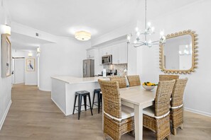 A modern kitchen with white cabinets, a central island, and a dining area with wicker chairs and a chandelier. - Dune it Right -Completely Updated with beach access, pool, hot tub, tennis, pickleball & gym (Santa Rosa Beach)