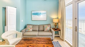 A room with a light blue wall, a sofa with floral cushions, a wicker chair, a wooden coffee table, and a view through a sliding glass door.