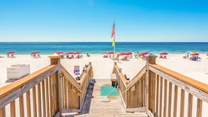 A wooden boardwalk leading to a beach with red umbrellas and a flag.