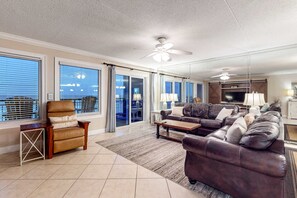 A spacious living room with a large sofa set, a wooden coffee table, and a chair. There are large windows with blinds, a ceiling fan, and a view of the outside. - Third-floor bright Gulf front condo with dreamy views - close to everything (Fort Walton Beach)