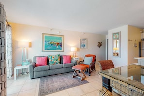 A living room with a grey sofa, colorful cushions, a glass coffee table, a wicker chair, and a small side table with a lamp. - Gulf-front condo with shared pool & gym, balcony, WiFi, & great view (Perdido Key)
