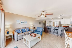 A living room with a sofa, coffee table, and a ceiling fan. There is a kitchen area with bar stools and a refrigerator. - Waterfront Gem - Shared Pool, Beach Access, Free WiFi, Washer/Dryer & Central AC (Fort Walton Beach)