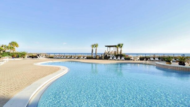 A clear blue swimming pool with a curved edge, surrounded by lounge chairs and palm trees, with a view of the sea in the background. - 13th-floor condo with amazing views, pools, gym & beach access (Perdido Key)