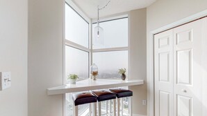 A kitchen with a white countertop, three bar stools, a hanging light fixture, and a view through the window. - 13th-floor condo with amazing views, pools, gym & beach access (Perdido Key)