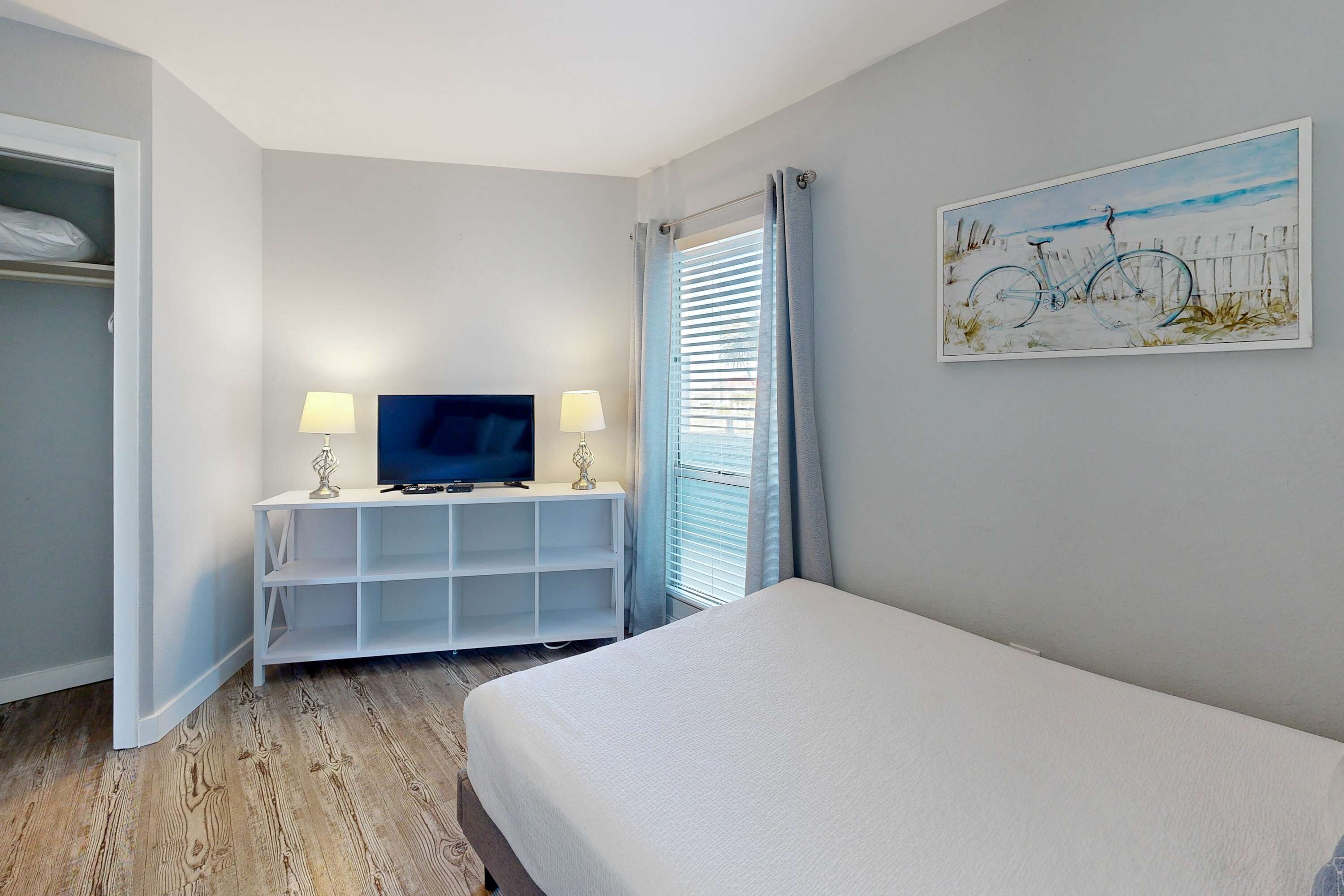 Une chambre à coucher avec un lit, un téléviseur posé sur un meuble blanc, un tableau accroché au mur et une fenêtre avec des rideaux.