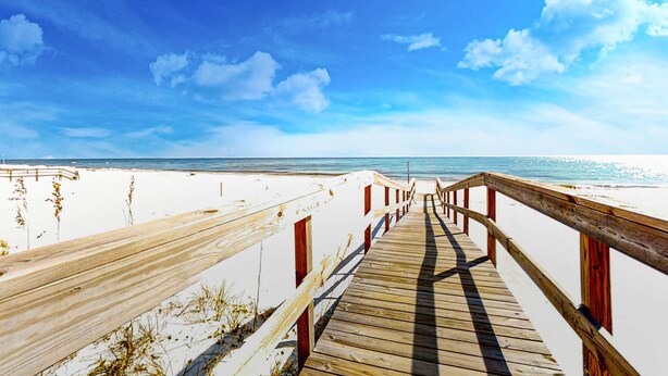 Ein Holzsteg führt zu einem Strand mit weißem Sand und wolkenlosem, blauem Himmel.