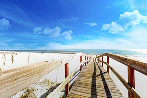 Ein Holzsteg führt zu einem Strand mit weißem Sand und wolkenlosem, blauem Himmel.