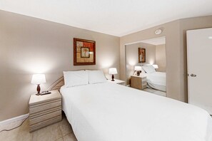 A hotel room with a large bed, two bedside tables with lamps, a nightstand, a framed picture on the wall, and a door. - Beachfront Condo w/ Gulf View, Balcony, WiFi & Shared Outdoor Pool/Gym (Perdido Key)