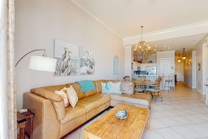 A spacious living room with a beige leather sofa, a wooden coffee table, and a dining area with a chandelier. - Open condo, on-site pool & hot tub, gulf-front balcony, on the beach (Perdido Key)