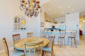 A dining area with a round wooden table and chairs, a chandelier, and a kitchen in the background. - Open condo, on-site pool & hot tub, gulf-front balcony, on the beach (Perdido Key)