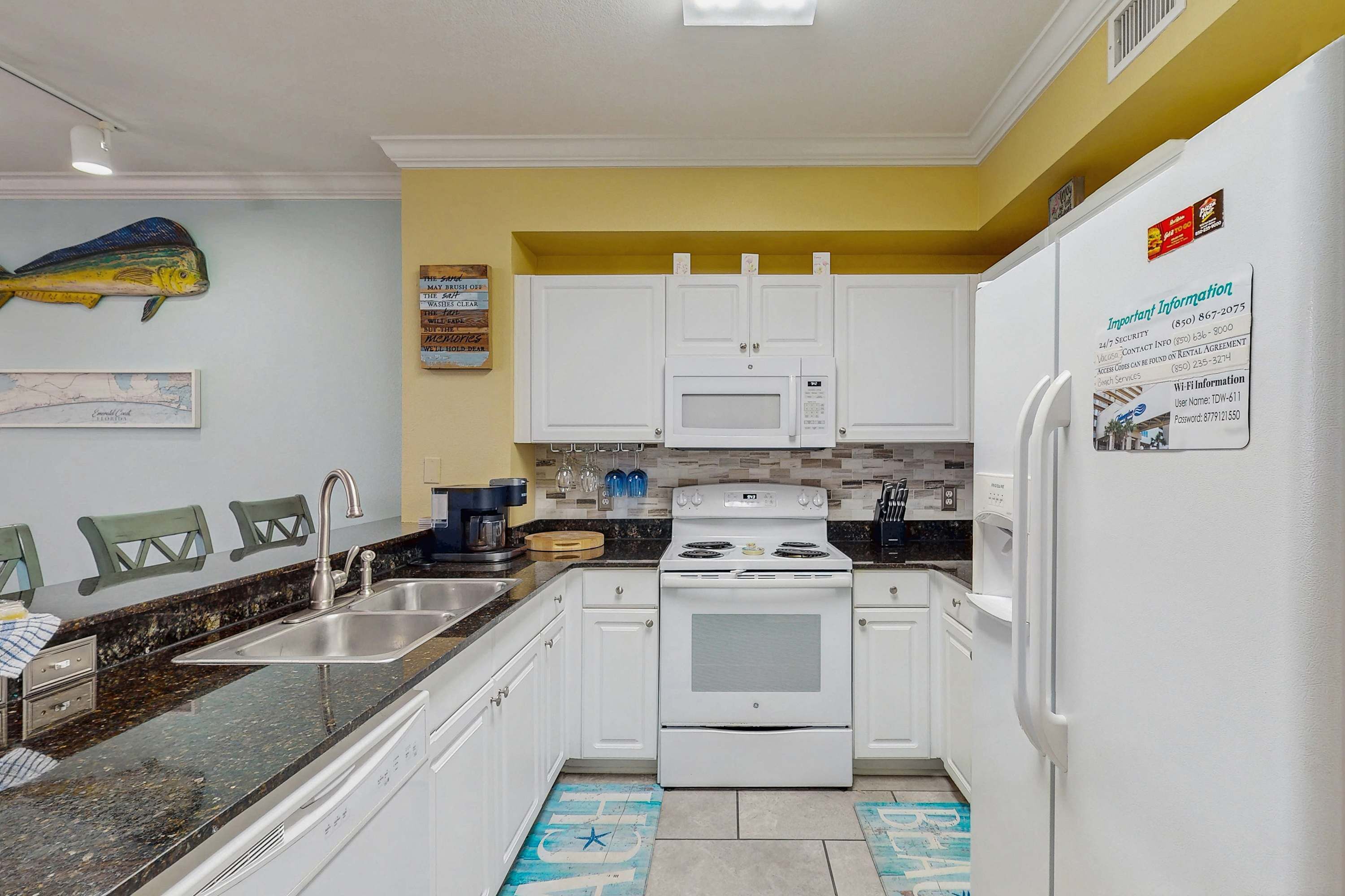 Una cocina compacta con gabinetes blancos, una estufa y un microondas. Hay un lavamanos con grifo, y un refrigerador a la derecha. Un mapa cuelga en la pared, y una decoración de pez está sobre los gabinetes.