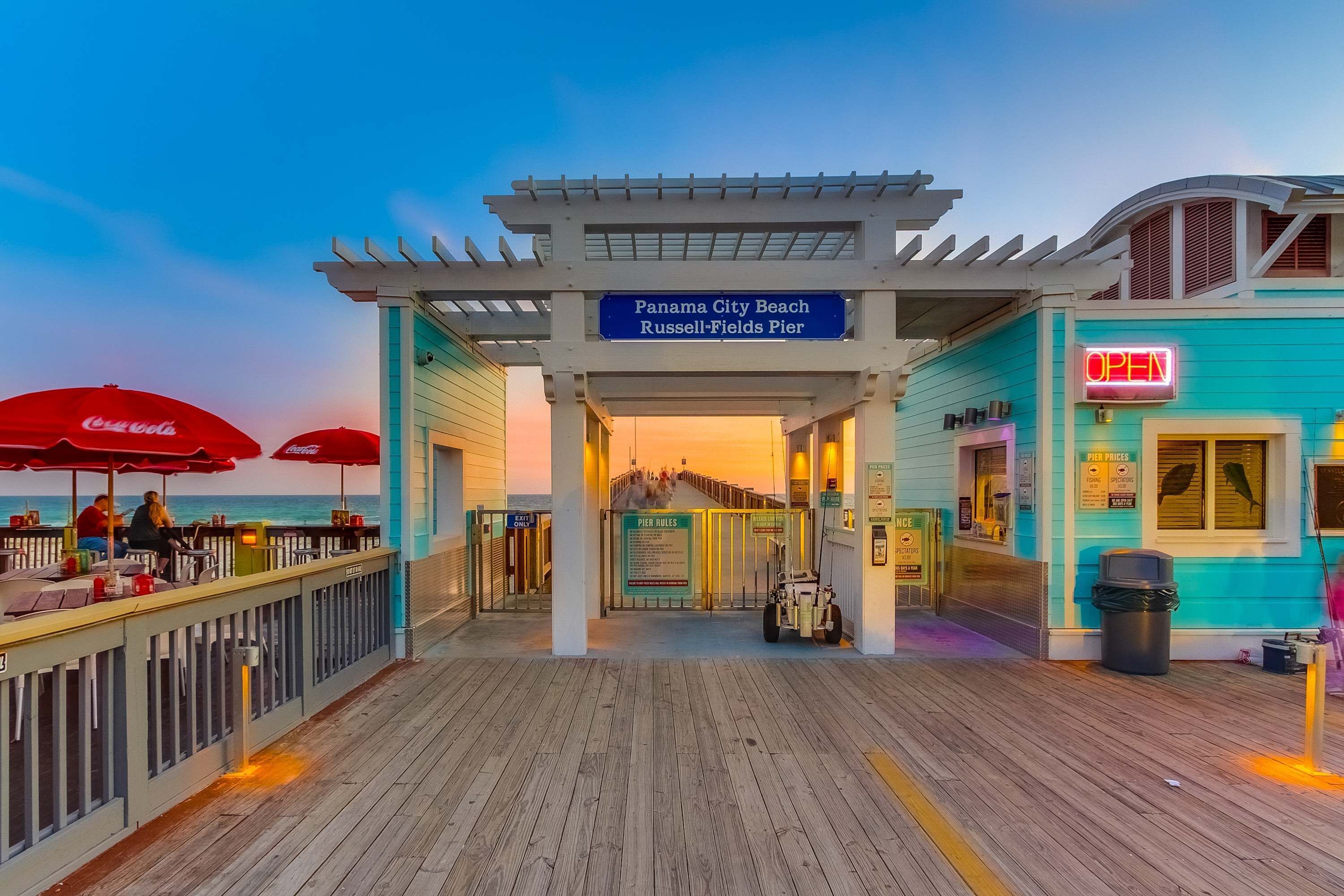 A pier entrance with a Coca-Cola umbrella, a sign for Panama City Beach, and a turquoise building with an 'OPEN' sign.