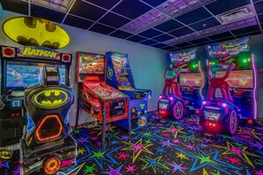 Une salle d’arcade lumineuse, avec différents jeux, dont un jeu sur le thème de Batman, et un sol aux motifs étoilés et aux couleurs vives.