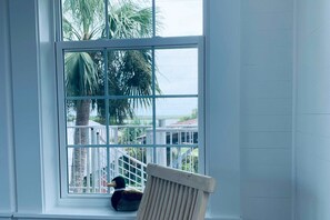 A room with a large window overlooking a white fence and palm trees. - Waterfront apartment with lovely views in walkable location (Apalachicola)