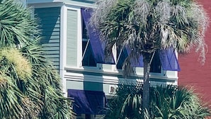 Un edificio con entrada en esquina, un balcón con contraventanas azules y una palmera en primer plano.