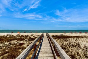 Ein hölzerner Steg führt zu einem Strand mit Sonnenschirmen und Sand.