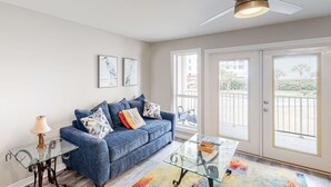 A living room with a blue sofa, glass coffee table, and a ceiling fan. - Updated condo with balcony, central AC, pools, hot tub, grills, & beach access (Perdido Key)