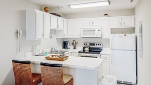 A modern kitchen with white cabinets, a microwave, stove, and a refrigerator. There is a dining area with wicker chairs and a small island with a basket. - Updated condo with balcony, central AC, pools, hot tub, grills, & beach access (Perdido Key)