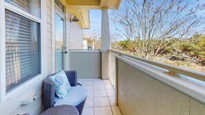 Uma varanda com sofá de vime, uma mesa, cortinas e vista para uma árvore e edifícios ao longe.